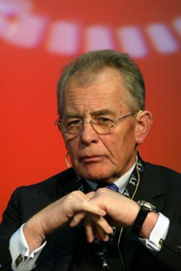Ein älterer Mann mit Brille und Anzug sitzt vor einem roten Hintergrund.