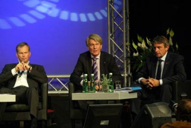 Drei Männer sitzen auf einer Bühne bei einer Podiumsdiskussion.
