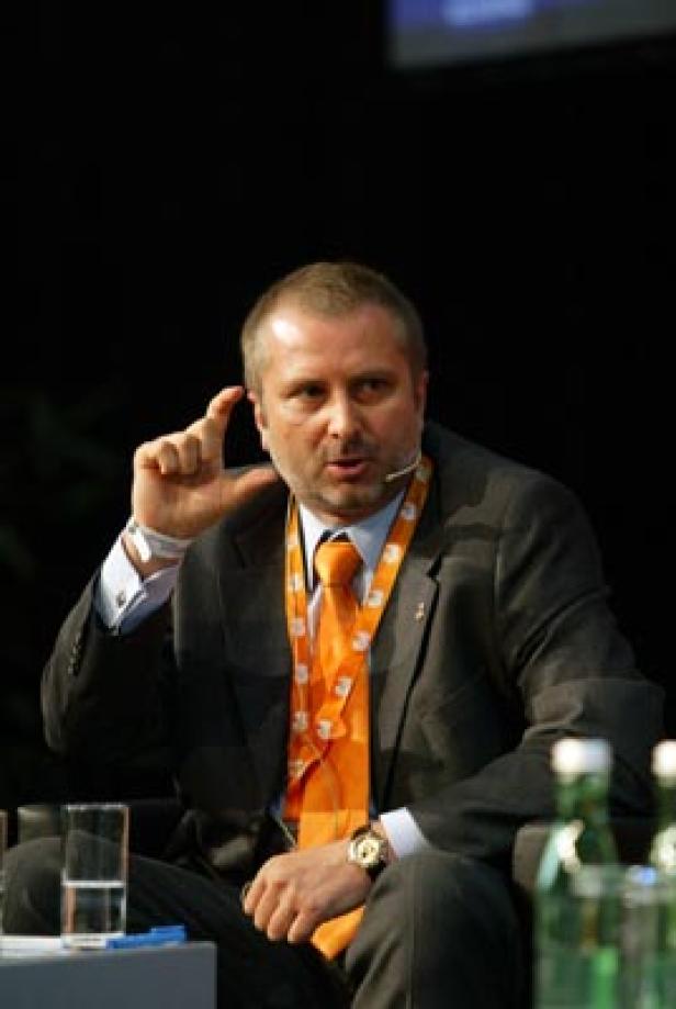 Ein Mann mit Anzug und orangefarbener Krawatte gestikuliert während einer Präsentation.
