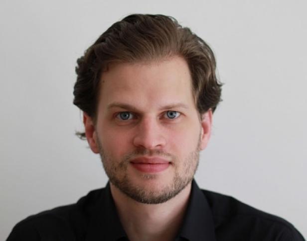 Porträt eines Mannes mit hellbraunen Haaren und blauen Augen vor einem weißen Hintergrund.