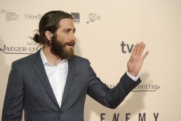 Jake Gyllenhaal auf einem Event, mit Anzug und Bart, die Hand erhoben.