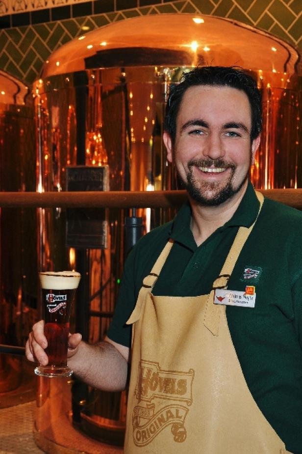 Ein lächelnder Mann im Hövels-Brauerei-Outfit präsentiert ein Glas Bier vor Kupferkesseln.