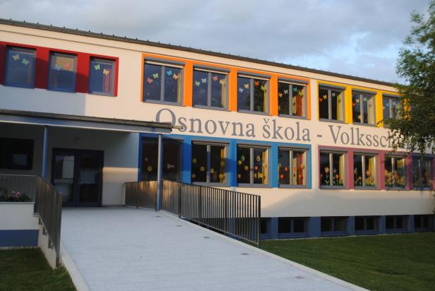 Die Fassade einer bunten Grundschule mit dem Schriftzug „Osnovna šola - Volksschule“.
