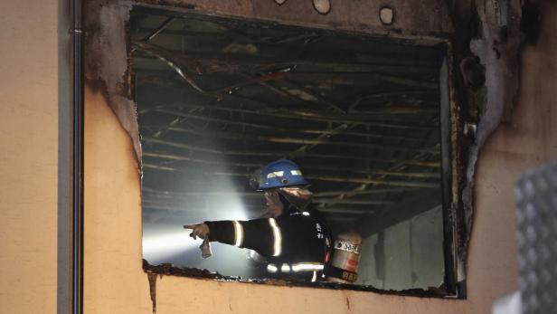 Ein Feuerwehrmann mit Helm und Sauerstoffflasche deutet in ein ausgebranntes Gebäude.