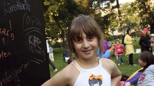 Ein junges Mädchen steht vor einer Tafel mit Kreidezeichnungen auf einer Wiese.