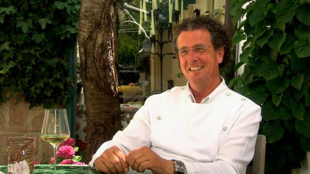 Ein lächelnder Mann in Kochkleidung sitzt im Freien mit einem Glas Wein.