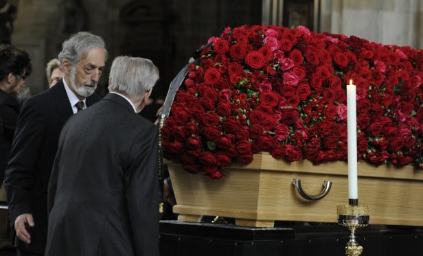 Zwei Männer stehen an einem Sarg, der mit roten Rosen bedeckt ist.