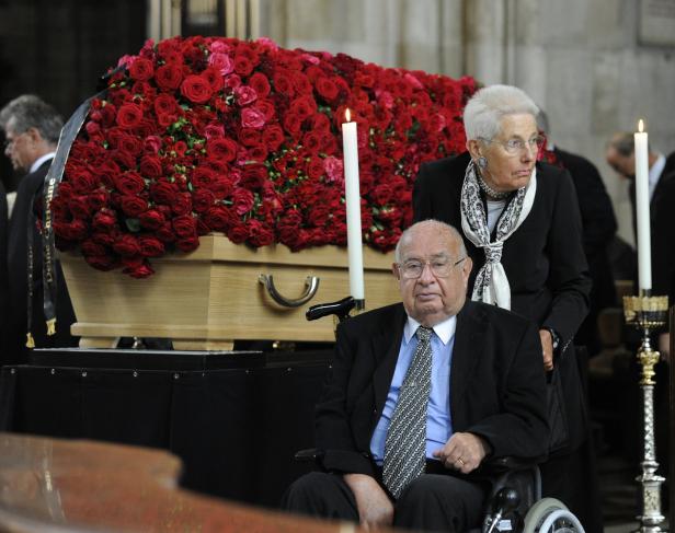 Ein Mann im Rollstuhl sitzt vor einem mit roten Rosen bedeckten Sarg.