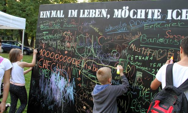 Kinder und Erwachsene schreiben Wünsche auf eine große Tafel mit der Aufschrift „Einmal im Leben möchte ich“.