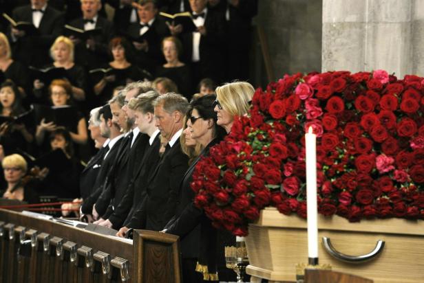 Trauergäste stehen neben einem mit roten Rosen geschmückten Sarg, während ein Chor singt.