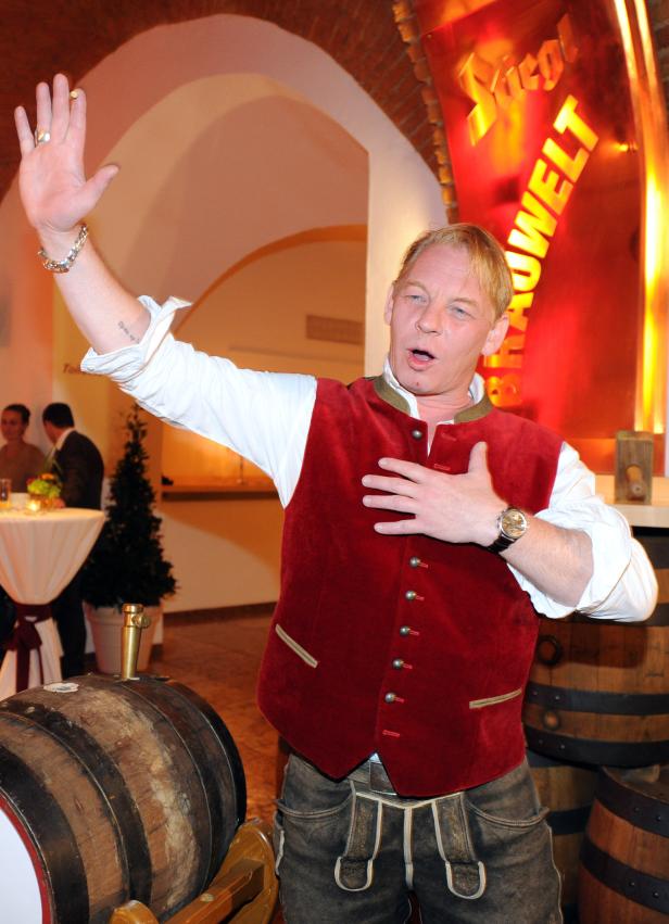 Ein Mann in Tracht steht vor Bierfässern und gestikuliert.