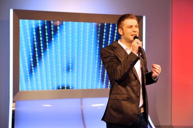 Ein Mann in einem Sakko spricht in ein Mikrofon vor einem Bildschirm.