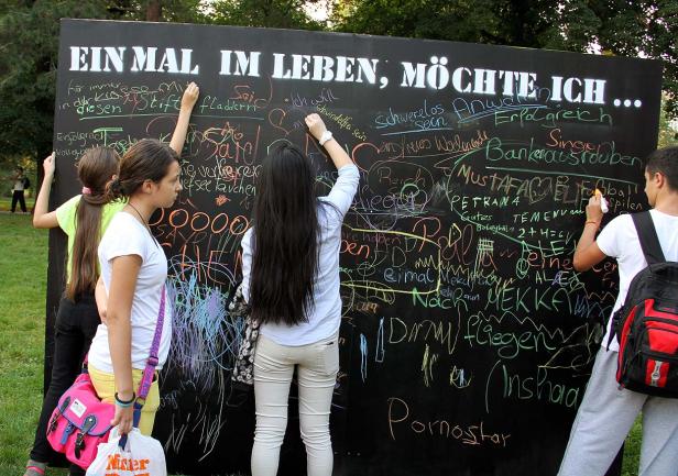 Mehrere Personen schreiben mit Kreide auf eine große Tafel mit der Aufschrift „Einmal im Leben möchte ich…“.