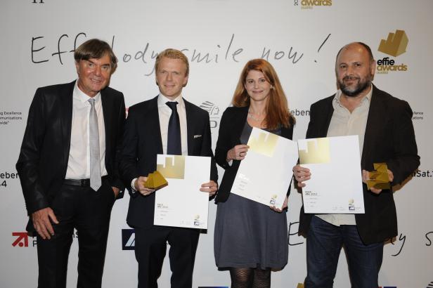 Vier Personen posieren mit goldenen Effie Awards und Urkunden vor einer Wand mit der Aufschrift „Everybody smile now!“.