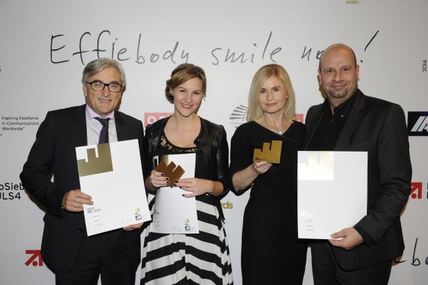 Vier Personen posieren mit goldenen Effie Awards vor einer Wand mit der Aufschrift „Effiebody smile now!“.