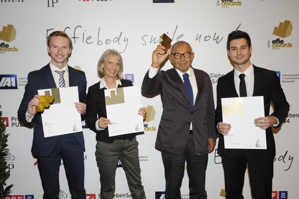 Vier Personen posieren mit goldenen Effie Awards-Statuen und Urkunden vor einer Wand mit Sponsorenlogos.