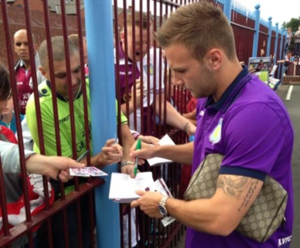 Ein Fußballspieler in lila Trikot gibt Autogramme hinter einem Zaun.