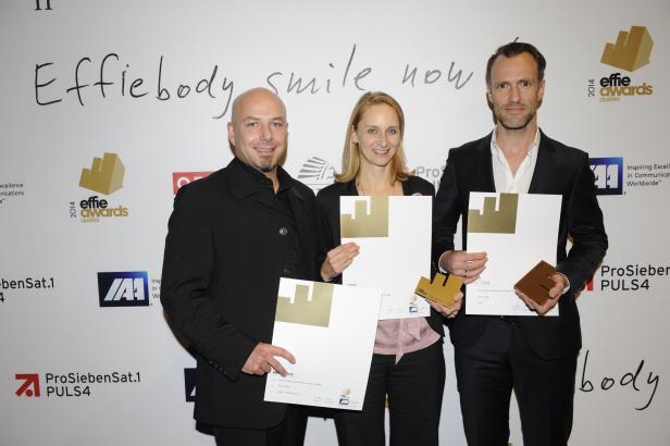 Drei Personen posieren mit Effie Awards vor einer Wand mit Sponsorenlogos.