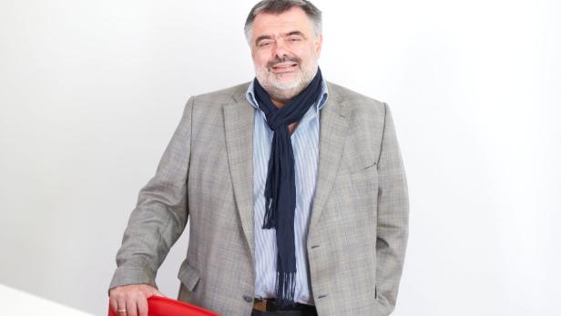 Ein Mann mit Bart, Sakko und Schal posiert vor einem weißen Hintergrund.