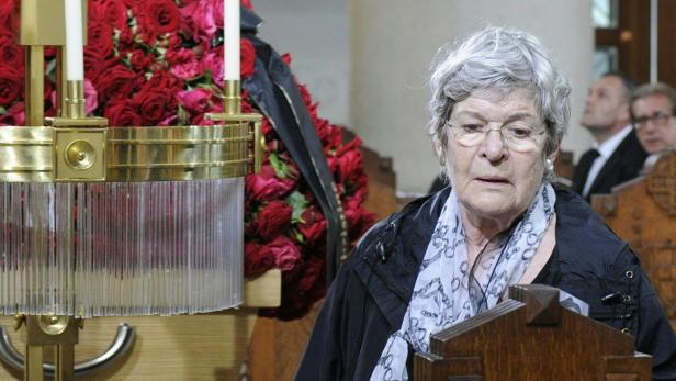 Eine ältere Frau steht an einem Rednerpult, im Hintergrund rote Rosen und Trauergäste.