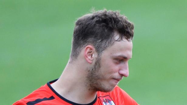 Ein Mann mit dunklen Haaren und einem roten Trikot blickt nach unten.