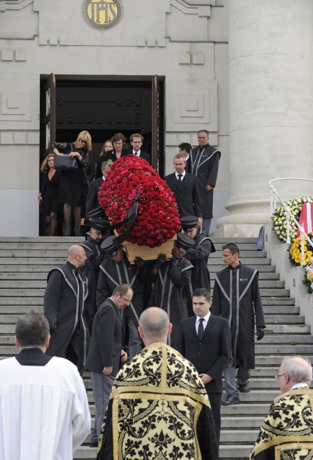 Ein Sarg, bedeckt mit roten Rosen, wird eine Treppe hinuntergetragen.