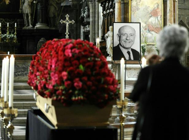 Ein Sarg mit einem großen roten Blumenarrangement in einer Kirche, daneben ein Porträt eines Mannes.