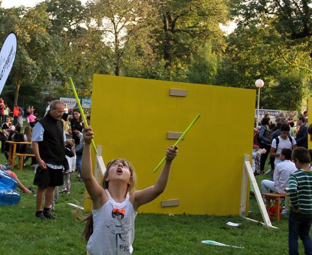 Ein Mädchen spielt mit Stäben vor einer gelben Wand auf einer Veranstaltung im Freien.