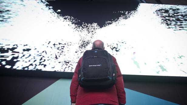 Ein Mann mit einem „DataStax“-Rucksack steht vor einer großen, hellen Leinwand.
