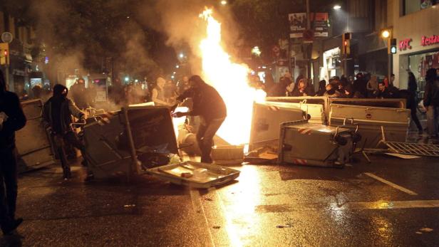Auf einer Straße brennt ein Feuer, umgeben von Müllcontainern und Menschen.