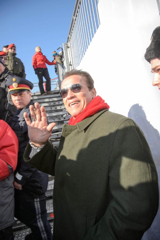 Arnold Schwarzenegger winkt in einem grünen Mantel und rotem Schal.