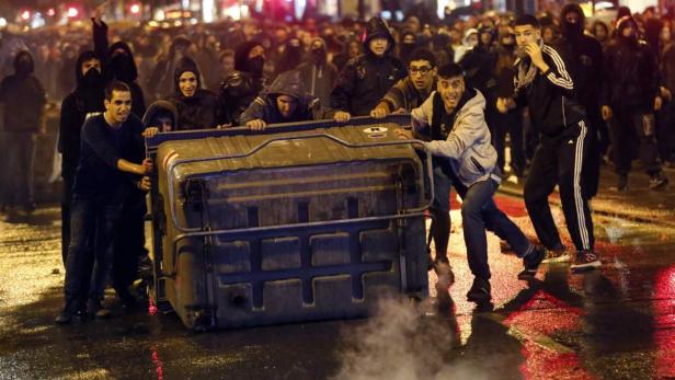 Eine Gruppe von Menschen schiebt einen umgekippten Müllcontainer durch eine dunkle Straße.
