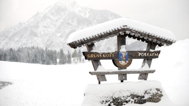 Ein schneebedecktes Schild mit der Aufschrift „Grüss Gott auf der Postalm“ vor einer Winterlandschaft.