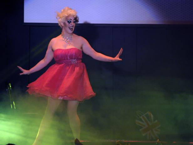 Eine Dragqueen in einem roten Kleid steht auf einer Bühne mit Union Jack im Hintergrund.