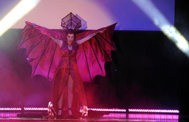 Eine Frau in einem roten Kleid mit Fledermausflügeln steht auf einer Bühne.