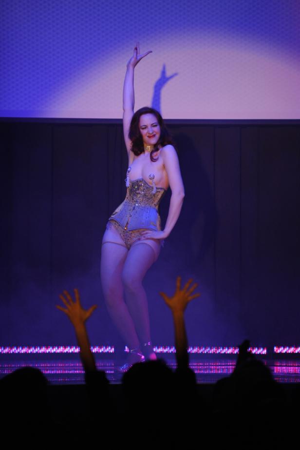 Eine Burlesque-Tänzerin in einem goldenen Korsett auf einer Bühne mit jubelndem Publikum.