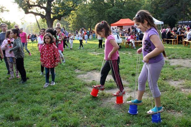 Zwei Mädchen laufen auf Dosenstelzen über eine Wiese, im Hintergrund eine Menschenmenge.