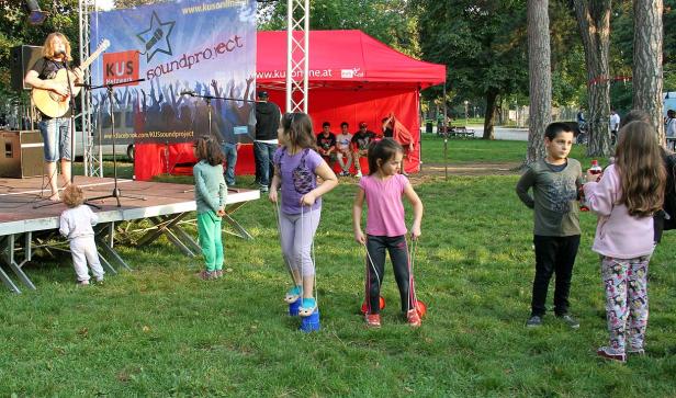 Eine Gitarristin spielt auf einer Bühne, während Kinder im Park spielen.