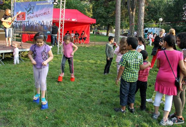 Kinder spielen auf einer Wiese mit Dosenstelzen, während im Hintergrund eine Band spielt.