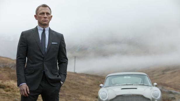 Daniel Craig als James Bond vor einem Aston Martin in einer nebligen Landschaft.