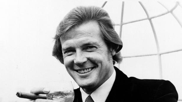 Roger Moore lächelt mit einem Drink und einer Zigarre in der Hand.