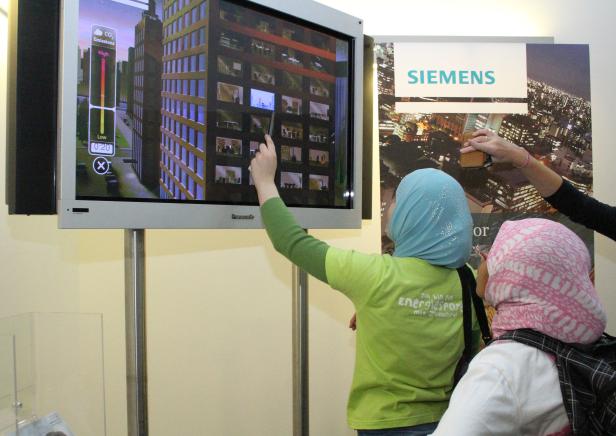 Zwei Frauen betrachten einen Bildschirm, der ein interaktives CO2-Emissions-Spiel von Siemens zeigt.