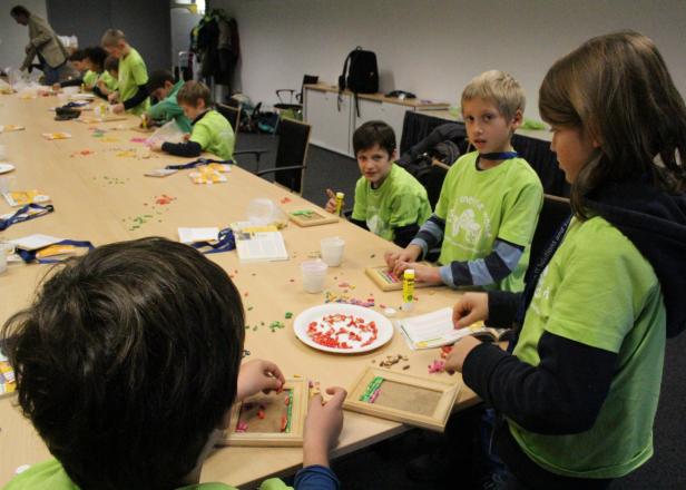 Eine Gruppe Kinder bastelt an einem langen Tisch mit bunten Perlen und kleinen Holzrahmen.