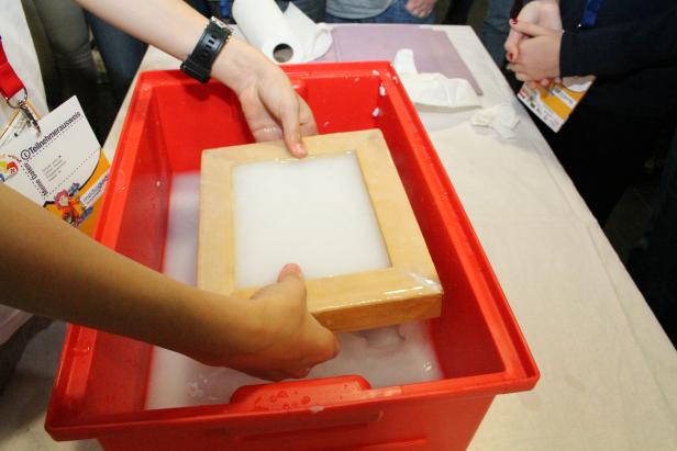 Eine Person schöpft mit einem Holzrahmen Papier aus einem roten Behälter.