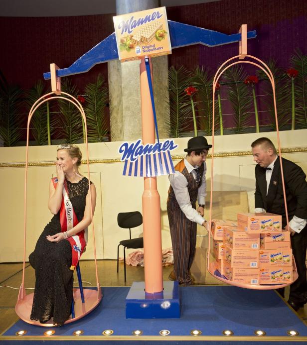 Eine Frau mit Schärpe sitzt auf einer Waagschale, während zwei Männer Manner-Waffelpackungen auf die andere Seite legen.