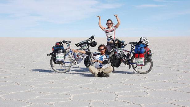 Ein Paar posiert mit ihren Fahrrädern auf einer weiten, hellen Ebene, möglicherweise einer Salzwüste.