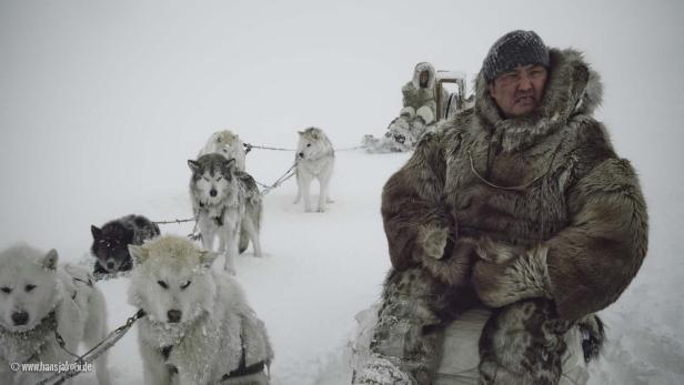 Ein Mann in Winterkleidung sitzt vor einem Hundeschlitten im Schnee.