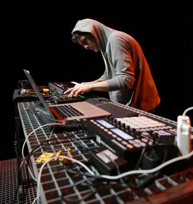 Ein Mann mit Kapuzenpullover bedient ein Mischpult und einen Laptop auf einer Bühne.
