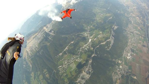 Zwei Wingsuit-Springer fliegen über eine grüne Landschaft mit Bergen.