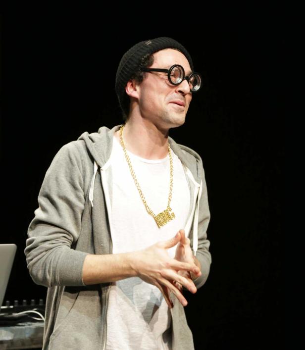 Ein Mann mit Brille, Mütze und Goldkette gestikuliert vor einem dunklen Hintergrund.